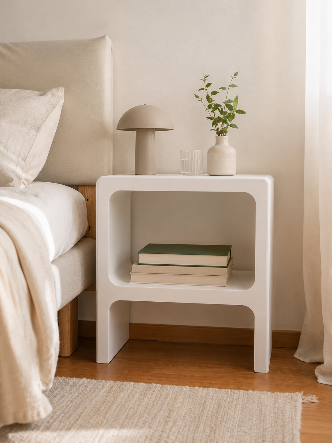 Mesa de cabeceira minimalista branca com cantos arredondados, candeeiro e livros, num quarto de estilo escandinavo