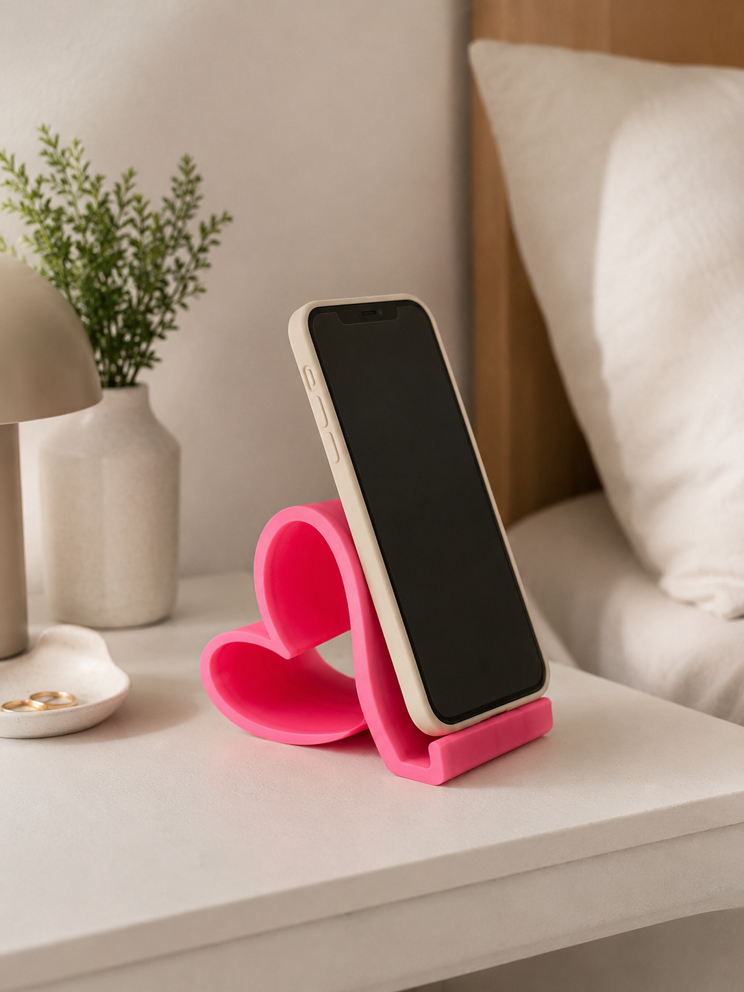 Suporte de telemóvel em forma de coração, cor rosa vibrante, com telemóvel apoiado numa mesa de cabeceira